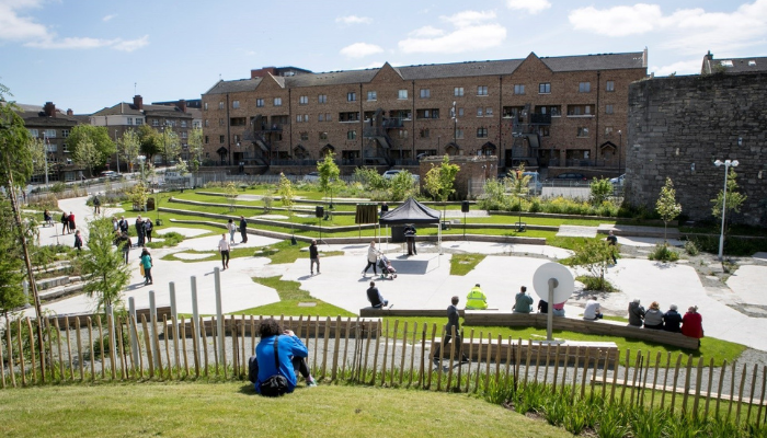 A widely shot image of a sunny communal square with much greenery around. 