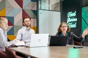 Three people sit in a meeting room with laptop looking at a screen