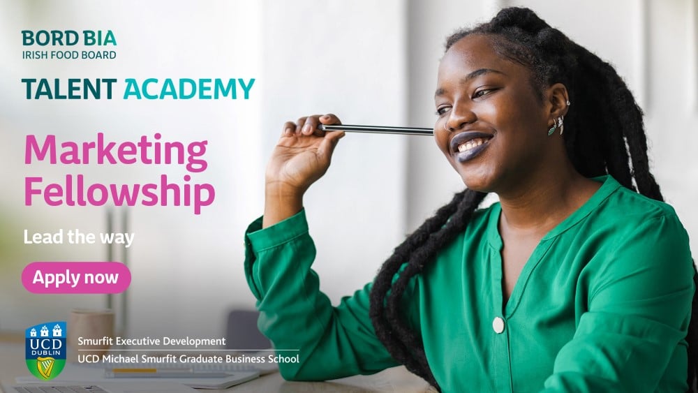 A promotional image for the Bord Bia Marketing Fellowship Programme. A women sits at a desk, holding a pen, smiling. On the left, text advertises the Marketing Fellowship Programme, with a slofan 'Lead the Way'.