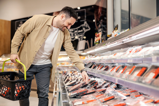 Man holding basket in supermarket while smiling, leaning over to pick up a packaged meat product.